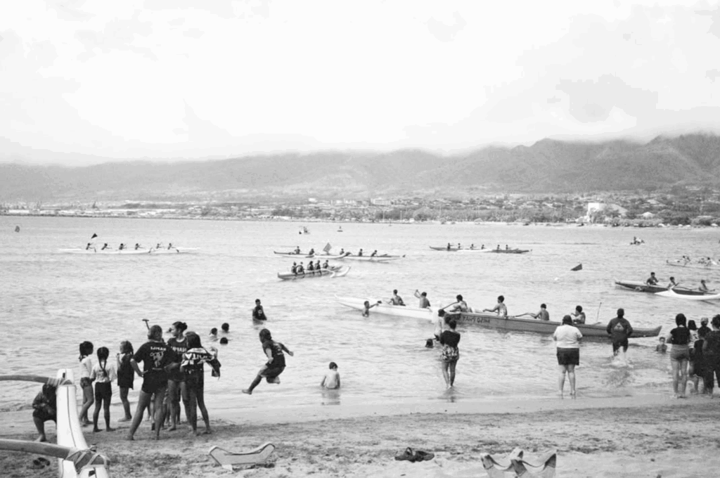 A black and white photo shows groups of people on a beach, some wading in the water, others watching outrigger canoes racing on the ocean. Mountains and a distant town are visible across the bay under a cloudy sky, while a few beachgoers relax with cups of Hawaiian coffee, savoring its rich aroma as they take in the lively scene.