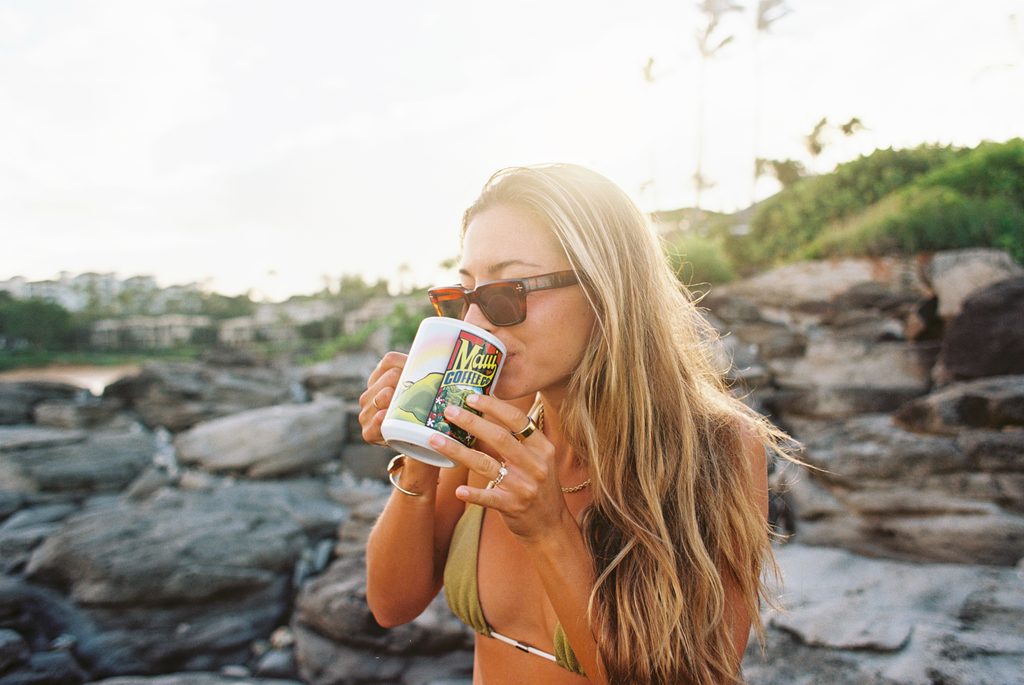 Woman pouring freshly brewed Maui Coffee Co. Hawaiian coffee into a mug beside the ocean in Maui, Hawaii, with a rainbow in the background.
