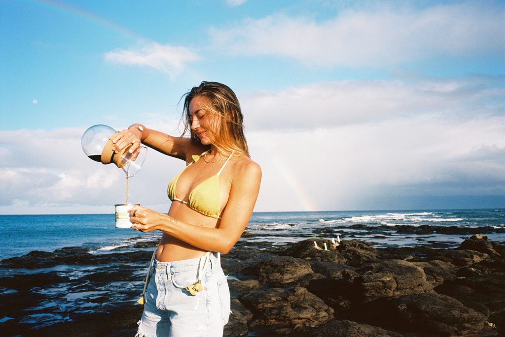 Woman pouring freshly brewed Maui Coffee Co. Hawaiian coffee into a mug beside the ocean in Maui, Hawaii, with a rainbow in the background.